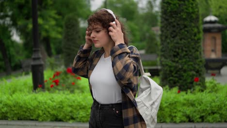 A-confident-brunette-girl-with-curly-hair-in-a-checkered-shirt-puts-on-white-wireless-headphones-during-her-walk-in-the-park-after-classes-at-the-university