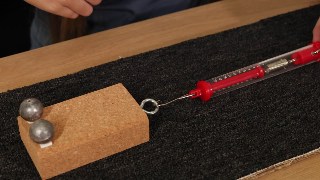 A hand uses a spring scale to measure friction by pulling a block with weights across a carpeted surface