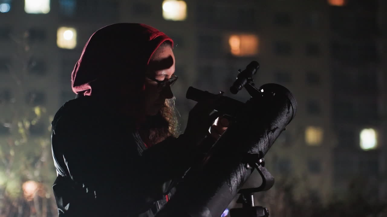 Appassionato di astronomia notturna sul tetto con telescopio puntato sul balcone urbano, figura incappucciata scruta attraverso l'oculare, bokeh di finestre cittadine sullo sfondo, silhouette con felpa rossa, paziente astronomo amatoriale che regola il telescopio