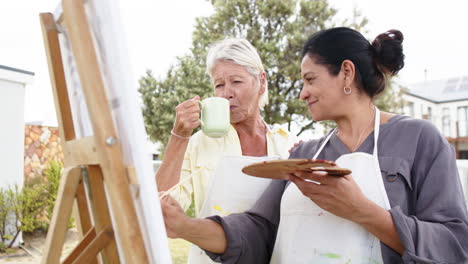 Zwei-Glückliche,-Vielfältige-Seniorinnen-Malen-Bilder-Und-Lächeln-Im-Sonnigen-Garten,-Zeitlupe