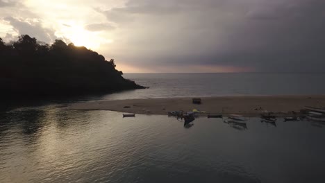 Rowboats-Docked-Along-The-Cape-Background-With-Beautiful-Sunset-Clouds-Partially-Hiding-Behind-Lush-Cliff-In-Rajbagh-Beach,-Canacona,-South-Goa,-India---Drone-Shot