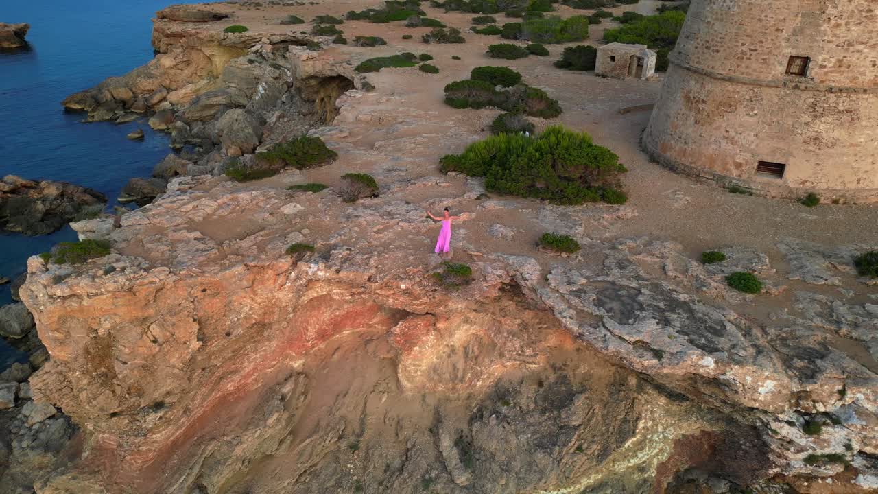 Donna spensierata in abito rosa a braccia aperte che si gode la libertà e una vista mozzafiato sull'isola di Ibiza vicino a Torre des Savinar al tramonto. Perfetto volo ascendente del drone per una vista aerea