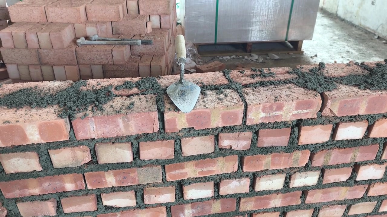 Double clay brick wall under construction on a job site with fresh wet mortar. A bricklaying trowel rests on top of the newly laid masonry line.