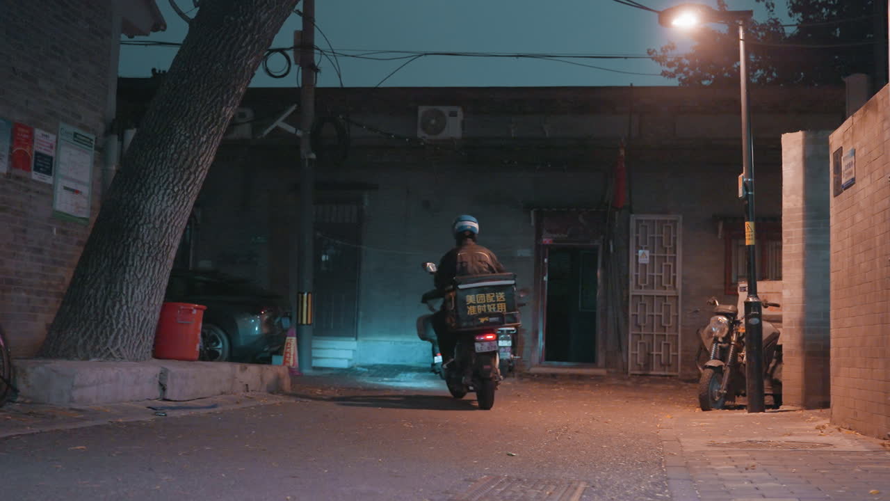 Scena di un vicolo vuoto, moto che parte da un portone, bauletto di consegna illuminato, motociclista con casco che si allontana sotto un lampione, ghiaia sparsa, figure sedute in lontananza su un uscio, atmosfera notturna cinematografica.