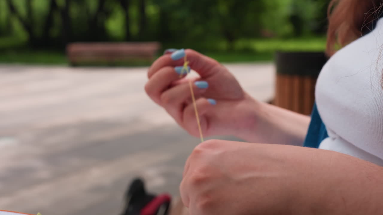 Close up hand view of person separating green thread with blurred park background, blue nails and steady fingers preparing embroidery outdoors, calm focus untangling floss under soft daylight