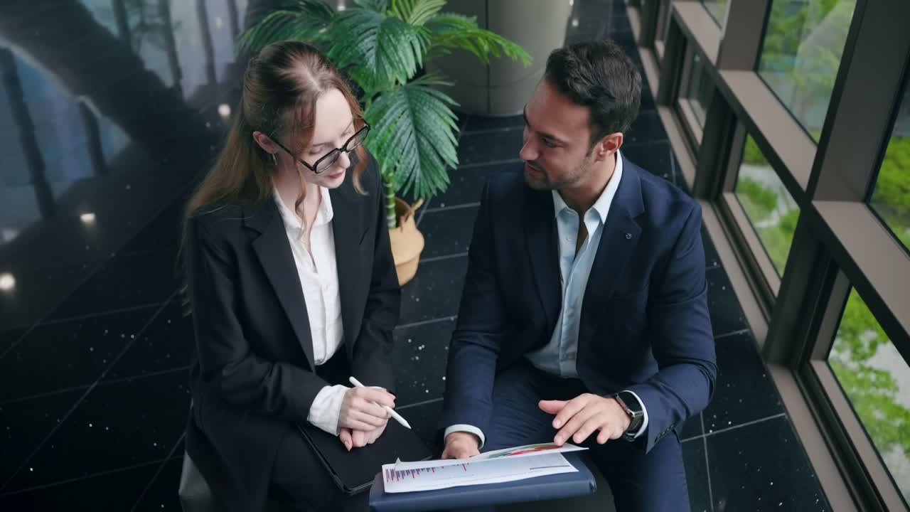 Due professionisti seduti vicino alla finestra che discutono di un documento con grafici e diagrammi, indicando con una penna - foto ad angolo elevato