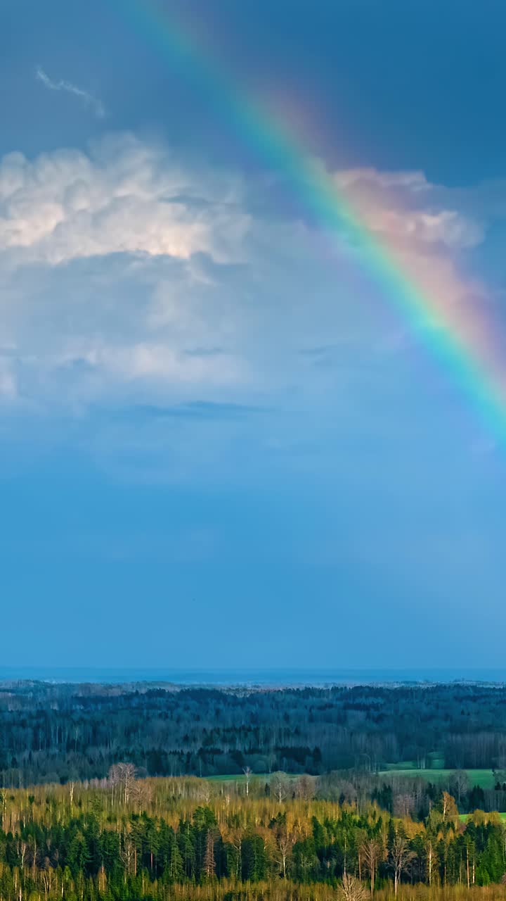 Vibrant rainbow arcs across dramatic blue sky above forested landscape, aerial hyperlapse