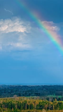 vibrant rainbow arcs across dramatic blue sky above forested landscape, aerial hyperlapse
