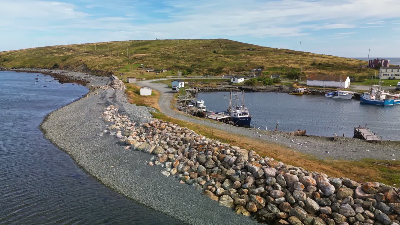 A drone flies over Ferryland’s small harbour, capturing a rocky breakwater protecting calm waters and a hillside community of white houses and cabins along Newfoundland’s rugged coast