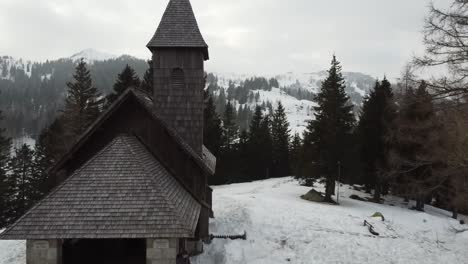 revealing shot old stone and wood church in winter forest with a traveller showing a ski resort with hotels and slopes