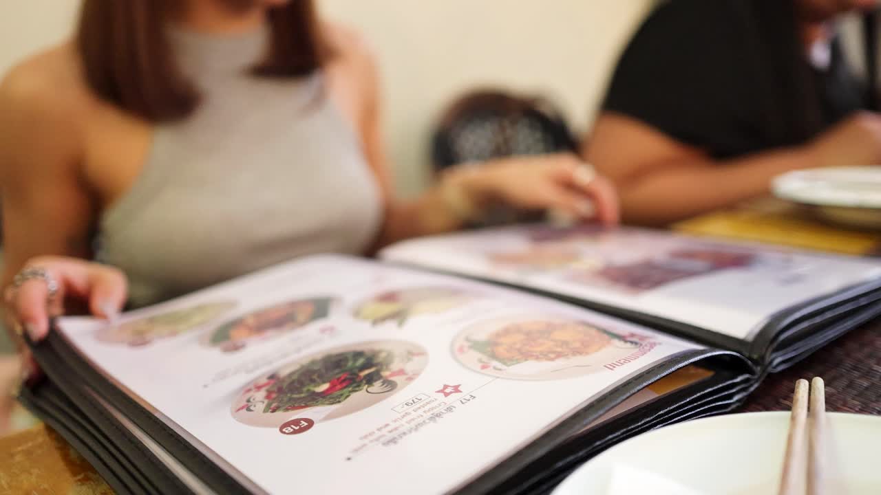 Two women sit at a restaurant table, flipping through a large illustrated menu of Northern Thai dishes in bright, natural lighting with shallow focus