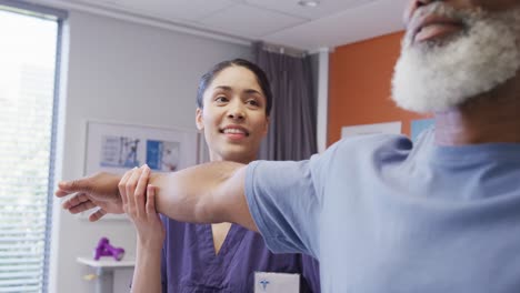 Happy-diverse-female-physiotherapist-and-senior-male-patient-working-on-arm-in-physical-therapy