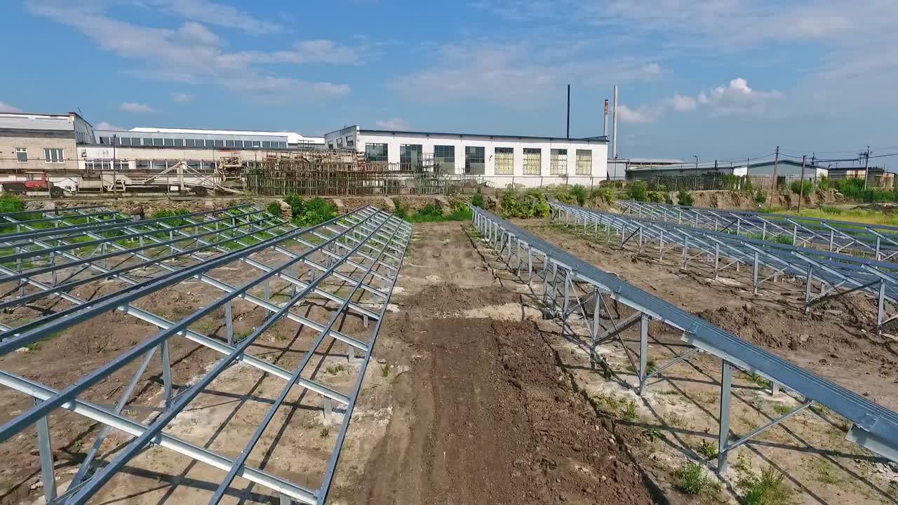 Installed metal supports on the field prepared for solar panels. Footage along the construction site with industrial zone at backdrop.
