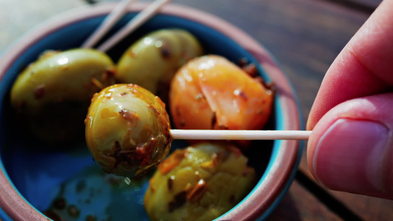 Close up of a hand picking up a green olive with a toothpick on a small bowl on a table