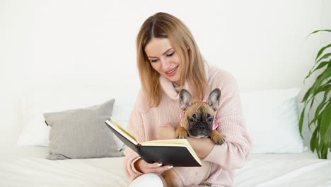 joven hermosa en un acogedor suéter rosa polvo y medias blancas se sienta en la cama con su perro y lee un libro