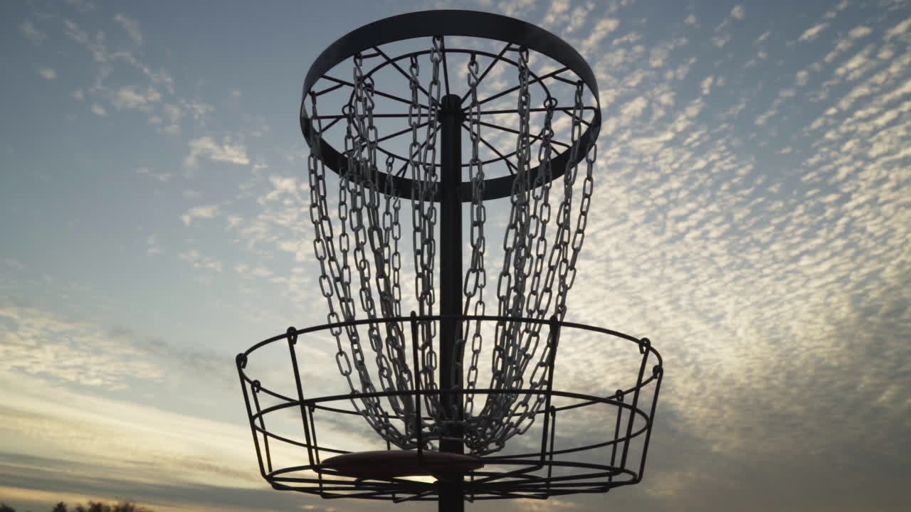 Il frisbee colpisce le catene centrali del cesto da disc golf sagomato contro il cielo nuvoloso al tramonto.