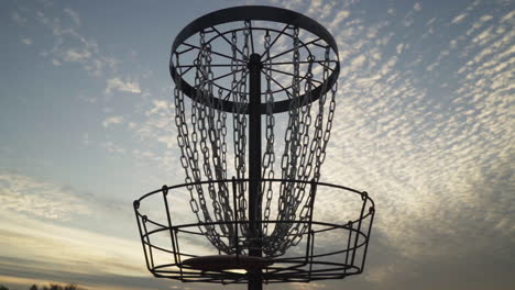 frisbee hits center chains of disc golf basket silhouetted by sunset cloud sky