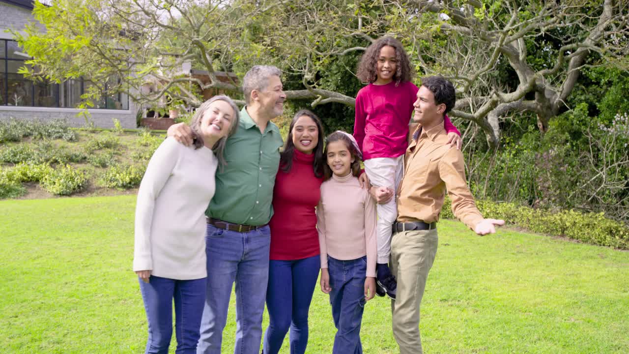 Family strolling across lawn under tree, kneeling and lifting child onto shoulders, posing portrait