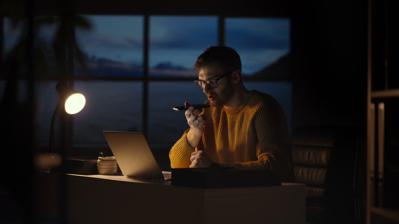 Focused young man lying on the sofa as he works on his laptop late into the night. Adult male working from home in living room at the night