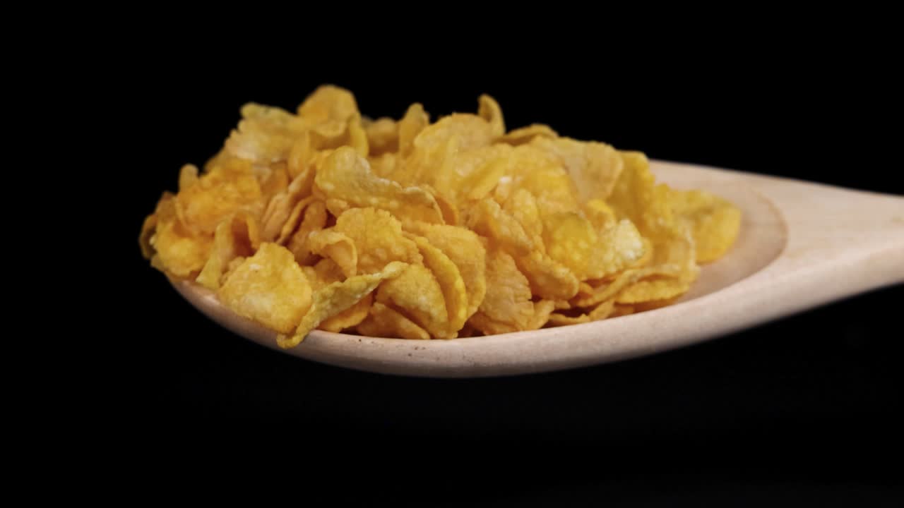 Golden cereal flakes cascade from a wooden spoon against a black background, highlighting texture and motion in slow motion