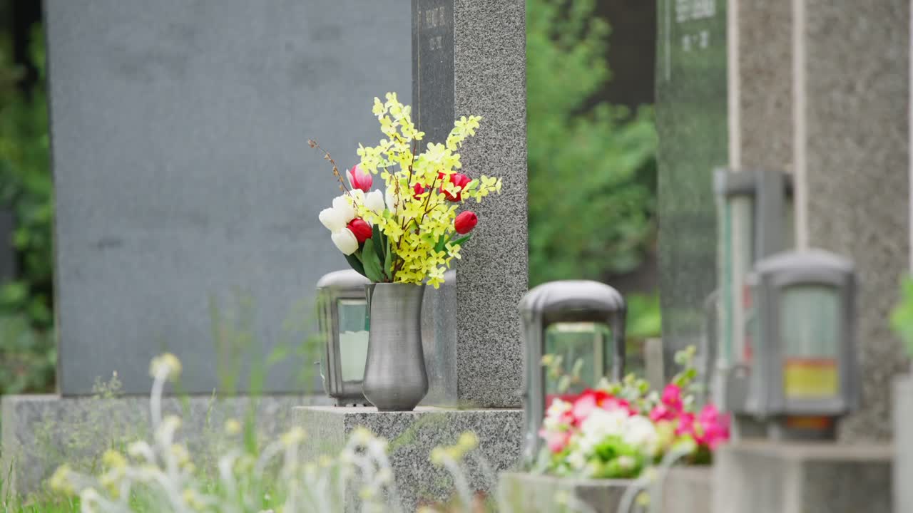 묘지 기념비에 놓인 꽃병 속 꽃들을 가까이에서 촬영한 사진입니다. 주변의 초록빛 식물도 보입니다. 부드러운 자연광으로 촬영하여 사색적인 분위기를 담아냈어요.