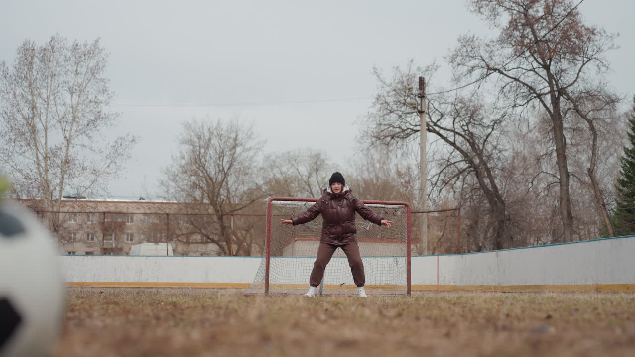 골키퍼 자세 예측, 집중하는 골키퍼가 있는 도시 축구 장면, 축구 선수가 슛을 하기 전 조용한 순간, 흐린 하늘과 텅 빈 나무들이 축구 연습을 위한 긴장된 분위기를 연출합니다.