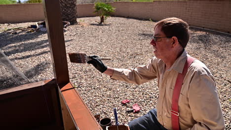 anciano sentado en el suelo mientras pinta un patio de madera en un día soleado