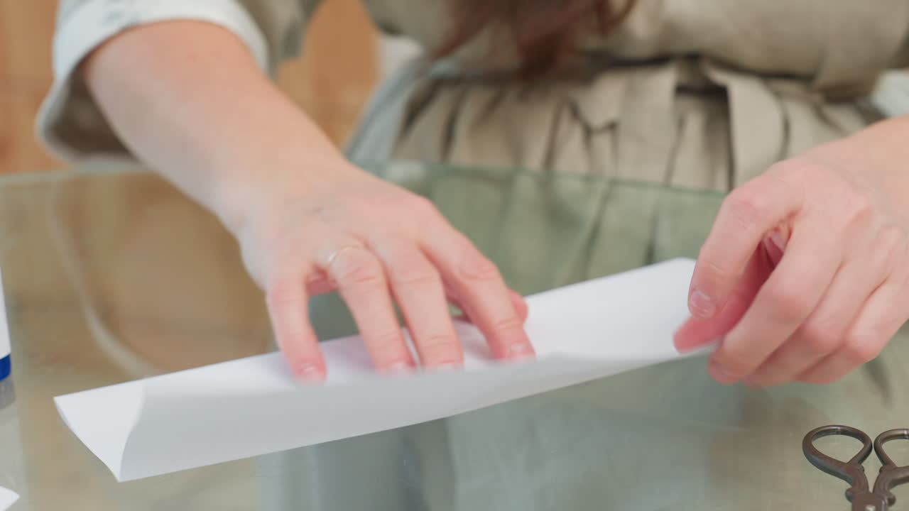 Close-up view of woman applying glue to join pieces of paper together on table with scissors placed beside it, focused on hands working on paper craft
