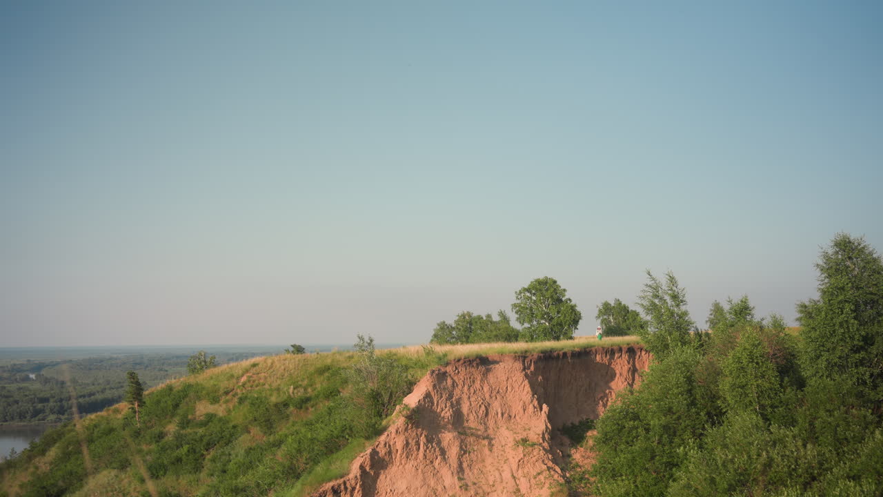 scenic view of rugged cliff edge surrounded by dense green trees and golden grassland under clear blue sky with flat distant horizon and tiny figure standing in background on warm sunny day