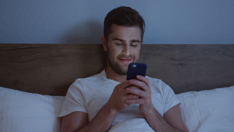 hombre caucásico acostado en la cama, charlando en el teléfono inteligente a altas horas de la noche antes de dormir