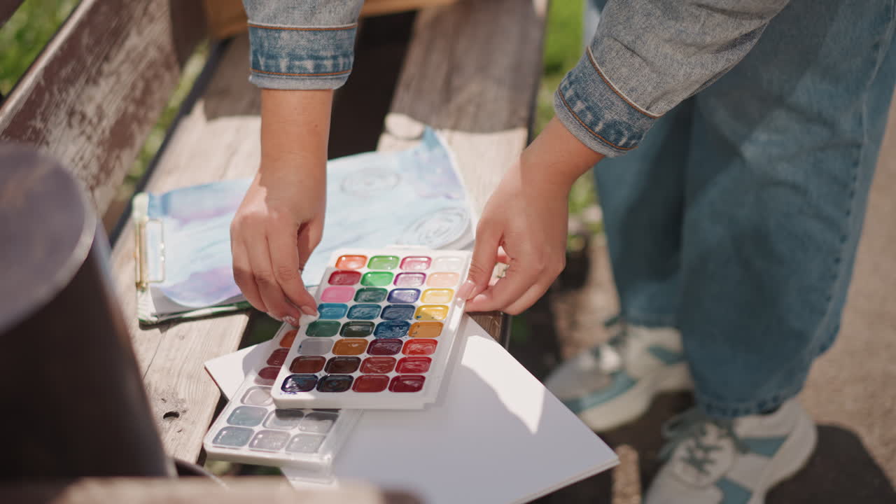 Outdoor-Aquarellpalette auf Bank, Künstler ordnet bunte Näpfchen an und bereitet Papier für Pleinair-Malerei vor, warmes Sonnenlicht, Jeansjacke und Turnschuhe sichtbar, Nahaufnahme Hände mischen Farbtöne und richten ein.