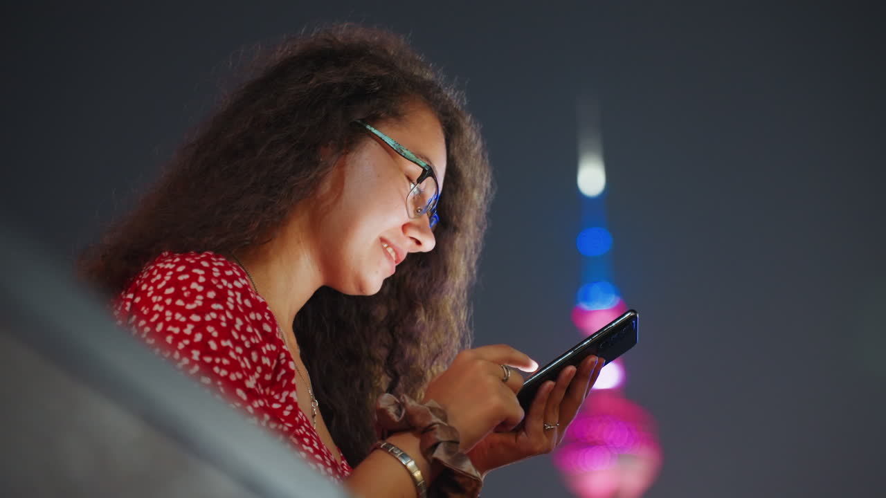Donna di notte vicino a una torre sorride mentre messaggia, giovane donna vicino a una torre illuminata controlla i messaggi e ride, la luce dello schermo evidenzia i capelli ricci e gli occhiali, vivido bokeh al neon, atmosfera energica di viaggio notturno.