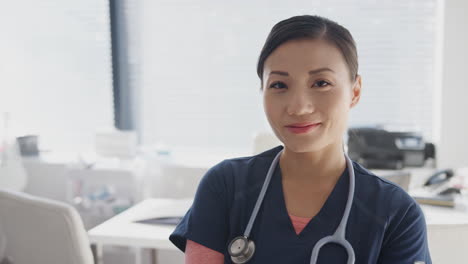 Portrait-Of-Smiling-Female-Doctor-Wearing-Scrubs-With-Stethoscope-Standing-By-Desk-In-Office