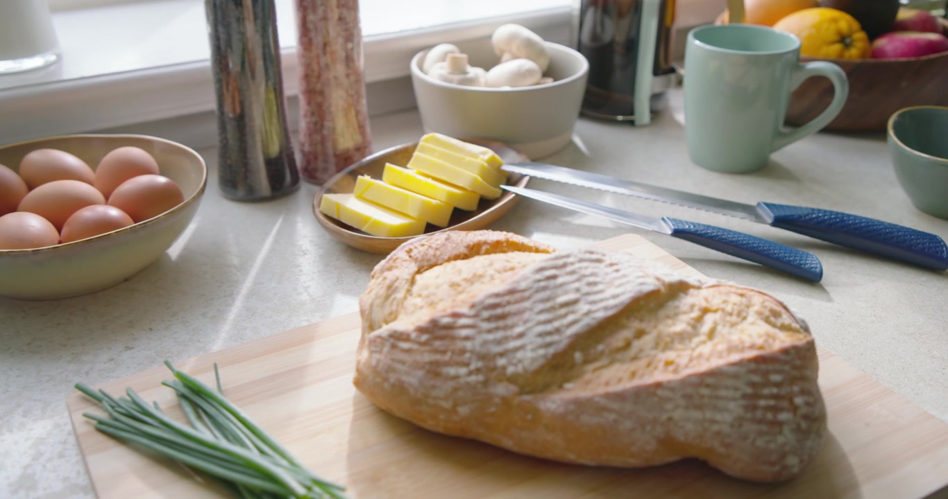 Camera is dollying by window, bringing crusty bread, butter and knives into focus for kitchen prep