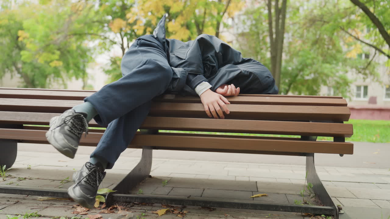 Estudante cansado descansando em um banco debaixo das árvores, jeans e jaqueta escura, com capuz, folhas de outono no chão, cena de parque tranquila, humor pensativo enquanto o estudante se prepara para o retorno às aulas.