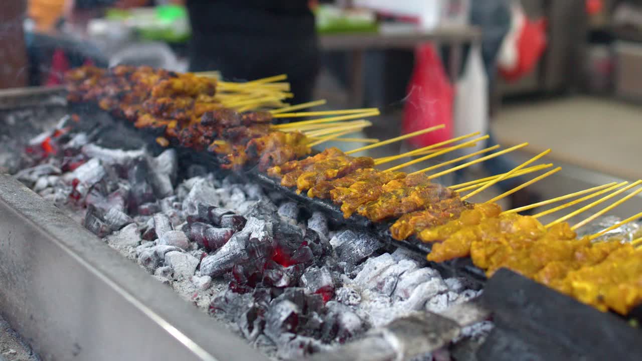 Jepretan jarak dekat tangan yang membalik sate daging marinasi di atas panggangan arang dengan asap.