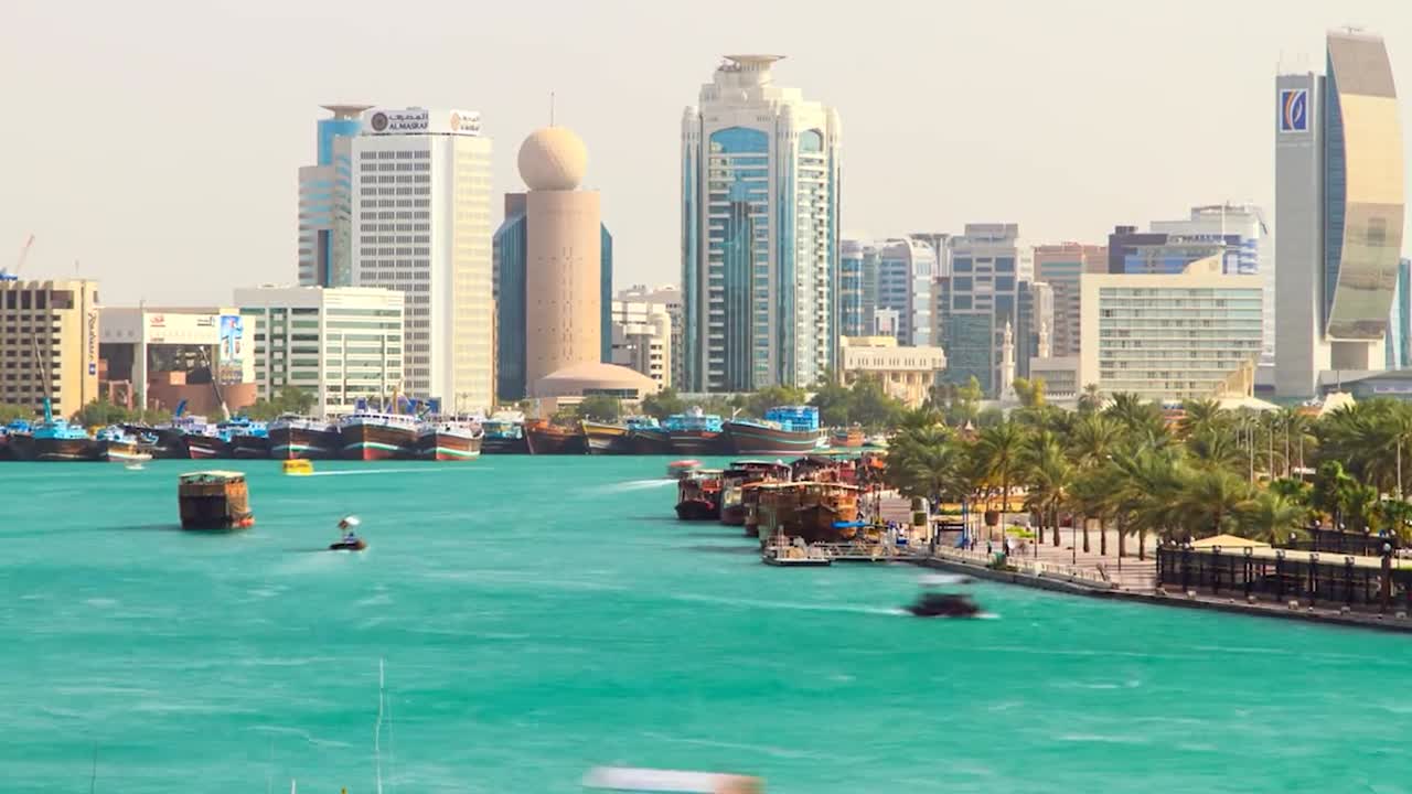 Time-lapse footage of Dubai Creek showcasing a vibrant waterfront cityscape. Traditional boats are docked along the turquoise shoreline, framed by palm trees and pedestrian walkways. Modern high-rise