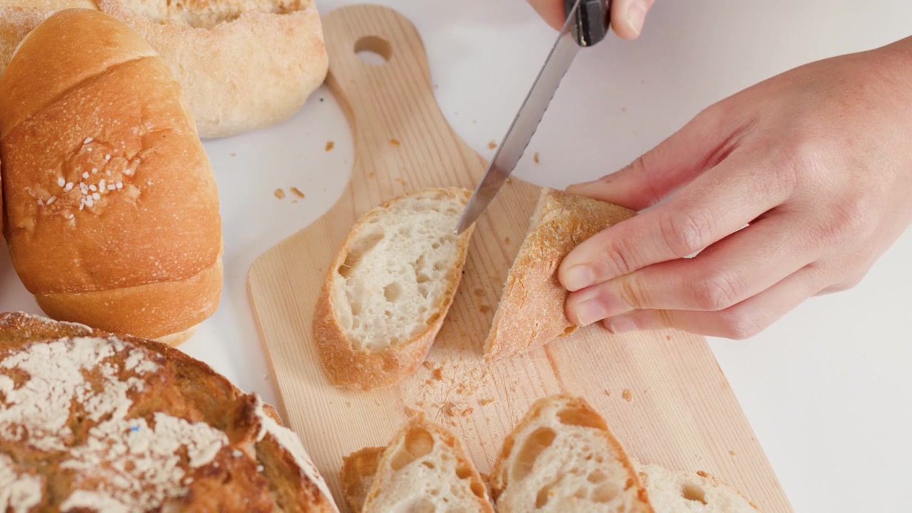 Hands use a serrated knife to slice crusty bread on a board with assorted loaves.
