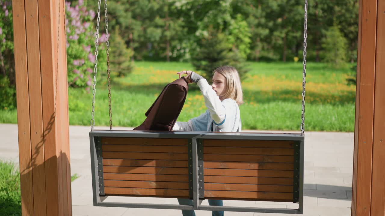 Una chitarrista bianca seduta su un'altalena che estrae la chitarra acustica dalla custodia, si sistema su una panchina di legno sotto la luce del sole in un parco verde. La musicista bionda controlla le corde e tiene delicatamente lo strumento, rilassata