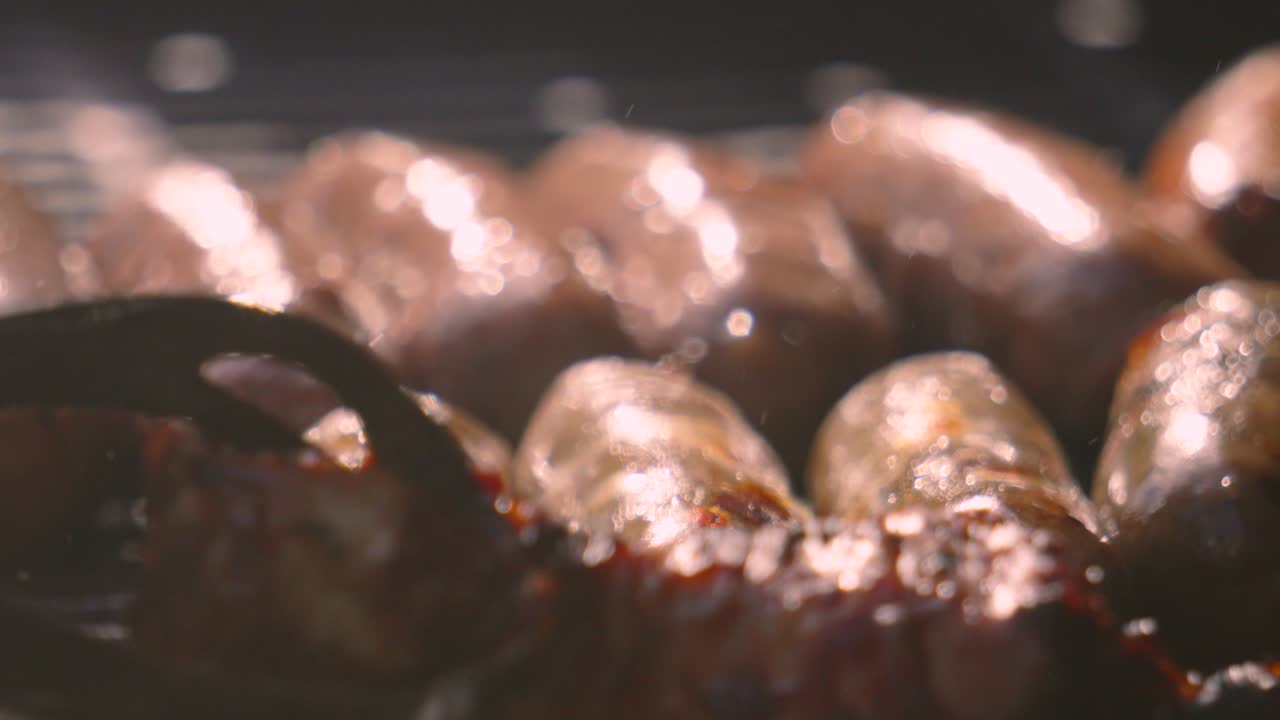 Close Up of Lots of Pork Sausages in a Row with Bright Sunlight Cooking Gently on BBQ with Heat Distortion with Oil Bubbling on Skin. High Fat Unhealthy but Delicious Meat Cooked Outside.