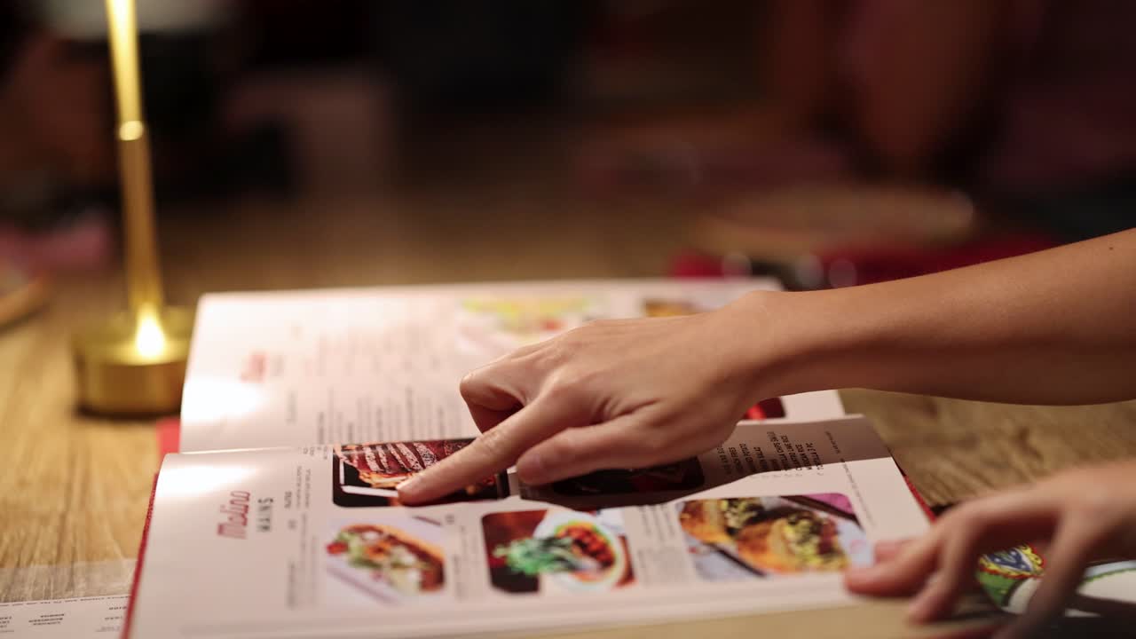 A hand flips through and points at dishes in a Mexican food menu under warm ambient lighting, shot on a wooden table in Bangkok, Thailand