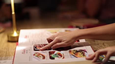 a hand flips through and points at dishes in a mexican food menu under warm ambient lighting, shot on a wooden table in bangkok, thailand