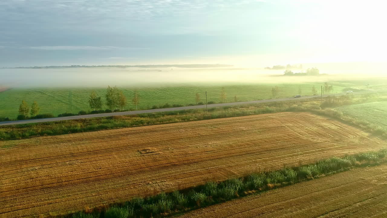 Aerial flyover rural fields with dew and clouds at road with cars during sunny day in the morning