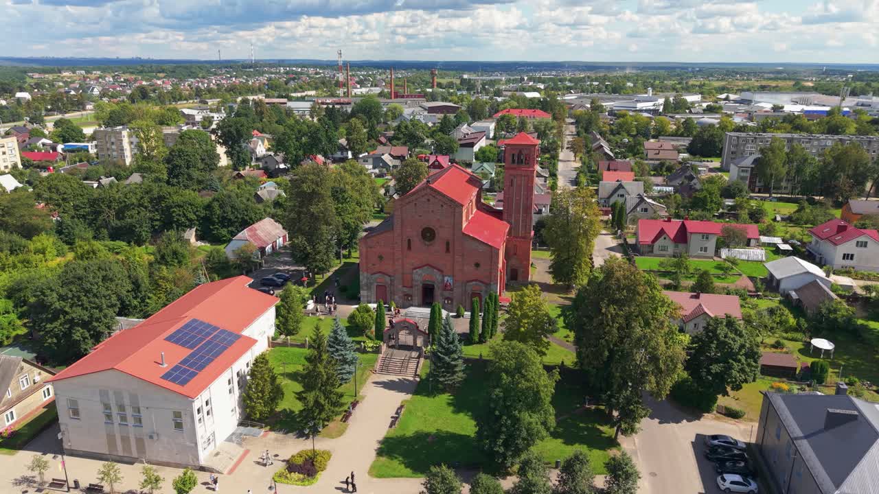 Aerial view of Lentvaris town in Lithuania featuring the prominent red brick Lentvaris Church surrounded by greenery, residential houses, and a bright summer sky