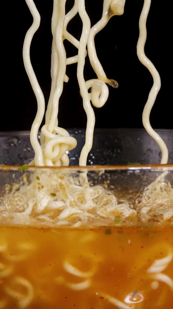 Noodles istantanei cotti vengono sollevati da una ciotola fumante di brodo arancione in un primo piano, con illuminazione drammatica e uno sfondo nero.