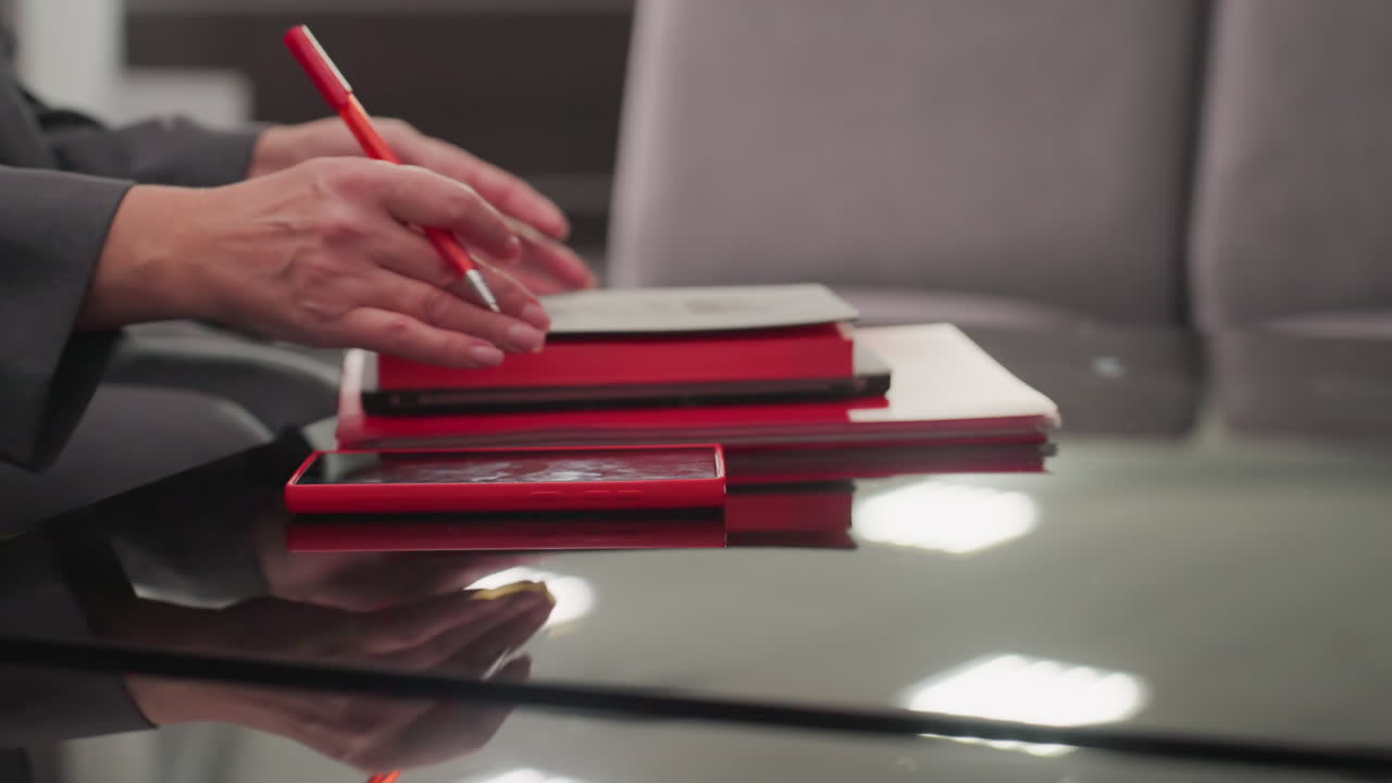 Primo piano della mano di una professionista che posiziona uno smartphone rosso sopra quaderni e documenti rossi in un ufficio aziendale. Una postazione di lavoro professionale con oggetti organizzati che riflette produttività e concentrazione.