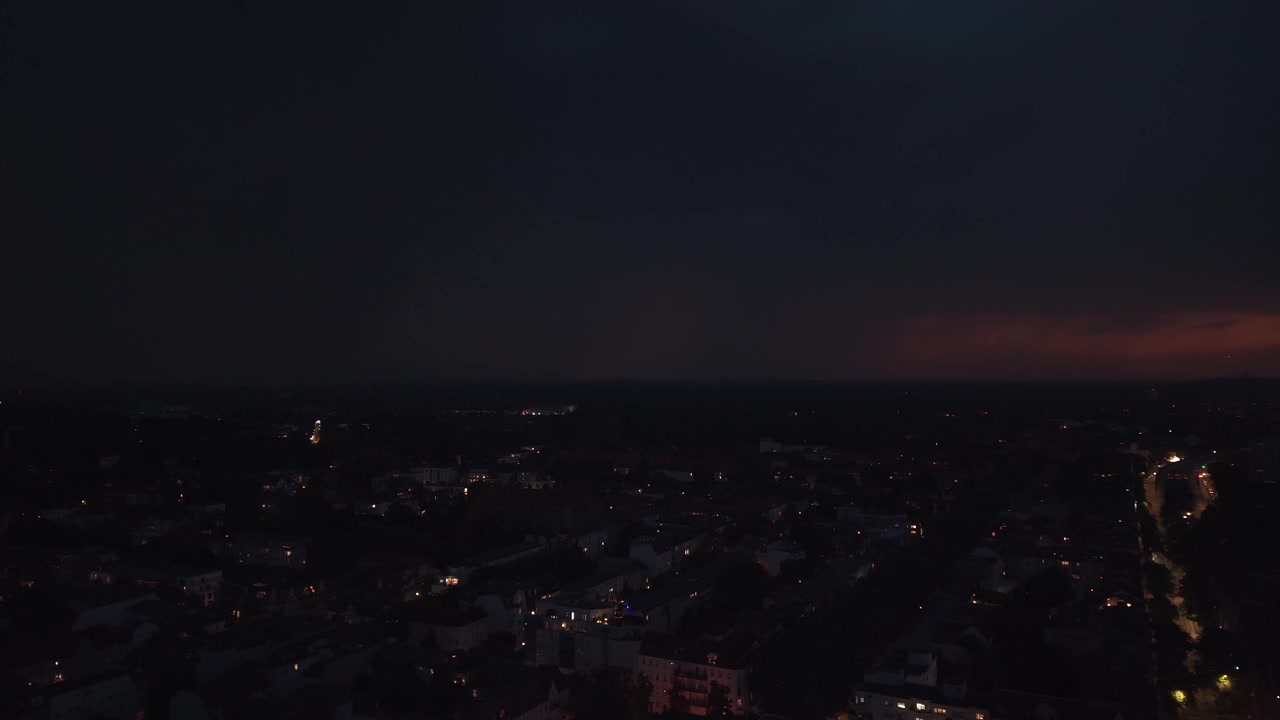 Blesky osvětlující temnou noční oblohu nad městskými budovami, zachycující dramatickou sílu letní bouře. Nádherný letecký panoramatický záběr stoupajícího dronu.