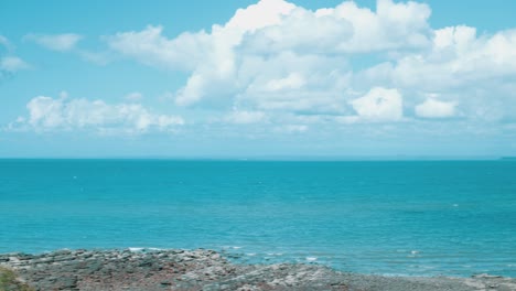 horizontal sweep of a beautiful, tropically blue beach that sits under a beautiful sky featuring white, fluffy clouds that float over the rocky, stone beach on a sunny day