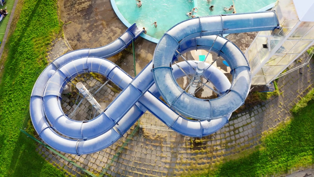 Una ripresa aerea dall'alto con drone mostra un grande scivolo acquatico blu a forma di otto accanto a una piscina pubblica con persone che nuotano e si divertono in una giornata di sole a Reykjavik, Islanda.