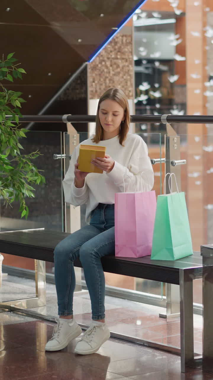 una mujer joven sentada en un banco en un centro comercial moderno y brillante recupera un libro amarillo de una bolsa de compras rosada, rodeada de bolsas de colores, barandillas de vidrio, plantas en macetas y un ambiente interior sereno